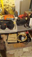 Orange power tools including a Skil saw, jig saw, black and orange oil changer, manual, and hoses, arranged on white plastic table and kitchen counters with stone basement wall background.