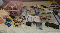 Overview photo showing large pink laundry basket with various toys inside and spread on bed, including diecast cars, action figures, and helicopter controller