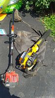 Two outdoor power tools on ground: yellow and black Poulan Pro blower and red and black Troy-Bilt weed trimmer.