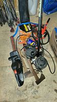 Photo showing an assortment of garden and hand tools including Black and Decker hedge trimmer, hand saw, hatchet, electric pumps, WD-40 can, and gloves on a wooden floor.
