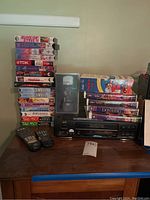 Overview of VHS player, remote controls and stack of VHS tapes on wooden cabinet.