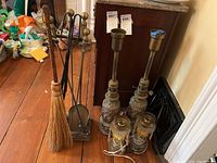 Two vintage lamps, metal fireplace tools with stand, and straw broom arranged on wooden floor