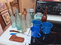 Overall lot showing six vintage bottles, three jars, three blue enamel mugs, and a folding cutter knife with brown leather sheath.