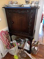Full front view of vintage wooden cabinet with closed single door and carved decorative trim, various holiday items around base, dishes on top not included.