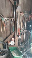 Photo showing an assortment of garden tools hanging on a wooden wall including rakes, shovels, and metal tools, with a green metal tub below containing materials.