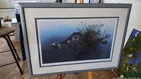 Framed limited edition print on stand showing full artwork of otter on log in water with plants, displayed indoors.