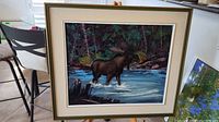 Framed oil painting showing full artwork of moose standing in river with forest background.