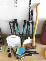 Photo showing hedge cutter, broom, axe, dustpan and green handled pruner against wall with other tools