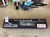Box of Mastercraft 2 ton cable hoist puller with product image and specifications, laid on a garage floor with other tools visible in the background.