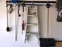 Step ladder and assorted car and garage cleaning tools hanging on wall including brushes, snow shovel, and hose head.