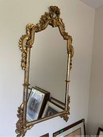Full view of the large wall mirror hung on a beige wall, showing the ornate gold carved frame with floral and scroll details.