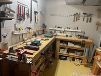 Wide view of workshop bench with assorted hand tools, clamps, hammers, screwdrivers, and saws on walls and bench surfaces.