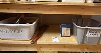 Wooden shelf holding plastic bins labeled for wrenches and drill bits with chisels, plus a large red pipe wrench on wooden surface.