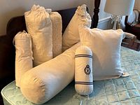 Seven white bed pillows and bolsters in various shapes including square, cylindrical, and rectangular crochet-covered bolsters arranged on bed mattress.
