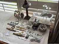 Image showing the antique Victorian oil lamp among multiple assorted decorative items placed on a window ledge with white shutter background. Items include angel figurines, brass drawer pulls, acrylic letter blocks, glass candlesticks, resin wreaths, and other small decor objects.