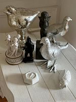 Photo showing the full lot arranged on white wooden table, highlighting three silver-tone bird figurines, black Cupid statue, white porcelain cherub music box, two small white porcelain decorative items, and soapstone carving.