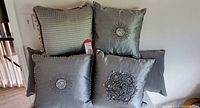 Six decorative accent pillows in grey, silver, and platinum tones displayed on furniture. Visible are various bead and sequin embellishments including sunburst and floral patterns, along with quilted texture pillows with frayed edges.