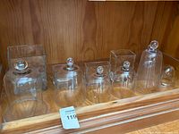 Photo showing six glass domes and three rectangular glass vases arranged on wooden shelf, clear glass with some rounded knobs on domes.