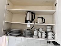 Shelved kitchen set showing stacked white ceramic plates and cups with black trim, along with a stainless steel electric kettle and glass coffee pot on upper shelves.