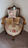 Full view of mid century wooden armchair with needlepoint seat and three floral needlepoint pillows arranged on the seat.