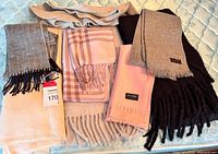 Lot of seven wool scarves showing various colors including pink, gray, cream, black and dark brown with fringes, folded and arranged on a mattress surface.