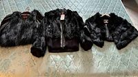 Three black rabbit fur jackets laid flat on a quilted surface. Left is a sleeveless vest, center is a zip-up jacket with ribbed cuffs and hem, right is a cropped fur jacket with short sleeves.
