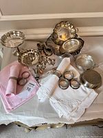 Silver plate serving baskets with ornate edges, pedestal compote dish, round and oval serving bowls, silver napkin rings, coasters, salt and pepper shakers, and pink and white embroidered linen napkins displayed on table.
