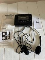 Photo of Sony Walkman FM/AM cassette player with attached wired headphones and user manual laid out on white red surface, showing front and top views.