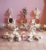 Five vintage metal wall candelabras painted white arranged on carpet, showing decorative floral and leaf motifs, two candle holders each, and small round mirrors