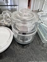 Close-up of vintage Pyrex Flameware coffee percolator showing glass body, metal band, handle, spout, and metal basket inside.