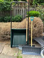Full view of the large green plastic snow mover with metal frame and two snow shovels leaning against it in an outdoor garden setting.