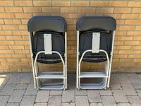 Two stacks of five folded Samsonite folding chairs each leaning against a brick wall, showing the folded backrests and frames.