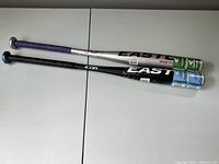 Two Easton youth bats laid horizontally. The top bat is silver and green purple grip, the bottom bat is black and blue with black grip.