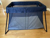 Blue Baby Bjorn Play Yard Light portable playpen set up on hardwood floor, showing mesh sides and metal legs.