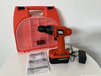 Orange and black Black & Decker cordless drill standing on battery with charger and manual in front of an orange carrying case with translucent front and Black & Decker logo.