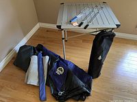 Camping lot with folded blue and gray tent, aluminum folding table with three roasting sticks on top, and tent carrying bag in corner