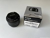 Canon EF 50mm f/1.8 II lens with attached front lens cap placed next to original box on white surface.