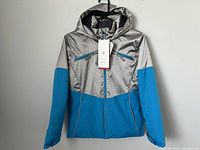 Front view of the Spyder Conquer youth ski jacket in silver and blue hung on a hanger.