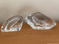 View of two Mats Jonasson lead crystal paperweights together, one shaped as a jumping salmon, the other as an iguana, both clear with frosted internal details.
