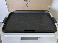 Top view of electric griddle with white base, black handles, and speckled non-stick cooking surface. Box in background.