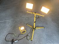Two halogen work lamps on yellow adjustable tripod stand and one smaller portable halogen lamp, all powered on showing bright light.