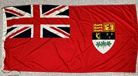 Front side of Canada Red Ensign flag showing the red field with Union Jack in the canton and detailed shield on the fly end with various heraldic symbols.