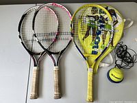 Three youth tennis rackets (two Babolat Genie 23 rackets and one yellow Wilson) laid out next to a yellow racket cover and a Tecnifibre training ball with attached rope on a table.