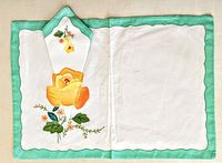 Full placemat with napkin folded and placed on top showing overall design and color scheme of yellow rose embroidery on white fabric with green border.