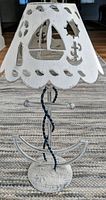 Front view of weathered white metal nautical tea light holder with cutouts of sailboat, anchor and other beach elements, showing twisted rope-like support and anchor base.