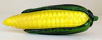 Side view of first Murano glass corn on the cob showing yellow kernels and green husk partially peeled back, on white background