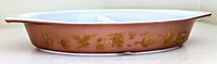 Side profile of the PYREX divided casserole dish showing brown exterior with gold Early American Heritage motifs and white interior separated by divider.