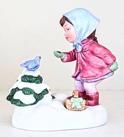 Front angle showing the girl in a pink coat, blue headscarf, feeding blue bird on snow-covered tree, with a basket on snowy base.
