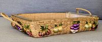 Side angle showing the wicker/rattan woven basket and embroidered multi-color fruits along the side with rope handles.