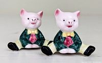Pair of vintage mid century ceramic pig salt and pepper shakers dressed in green jackets, red ties, and yellow bows, seated with legs spread apart.